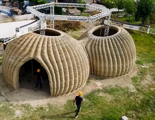 Two large nearly spherical structures stand on the ground with a person walking by them. They are more than twice as tall as the person. Above the structures, a metal structure is suspended in the air with additional machinery connected to them.