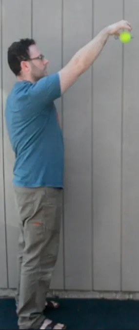 A person in a blue shirt and khaki pants stands sideways against a gray wall, holding a yellow tennis ball above their head with an outstretched arm.