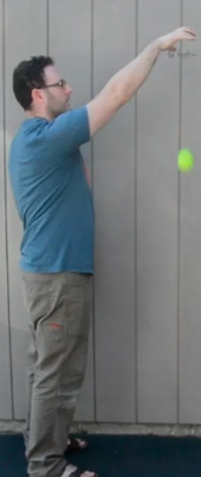 A man in a blue shirt and khaki pants stands sideways  against a gray paneled wall, arm extended upward, and has dropped a yellow tennis ball. The ball is approximately chest height of the man.