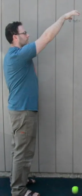 A man in a blue shirt and khaki pants stands sideways  against a gray paneled wall, arm extended upward, and has dropped a yellow tennis ball. The ball is on the ground.