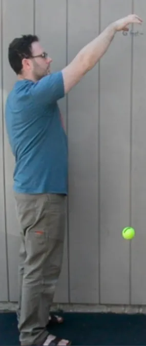 A man in a blue shirt and khaki pants stands sideways  against a gray paneled wall, arm extended upward, and has dropped a yellow tennis ball. The ball is approximately knee height of the man..