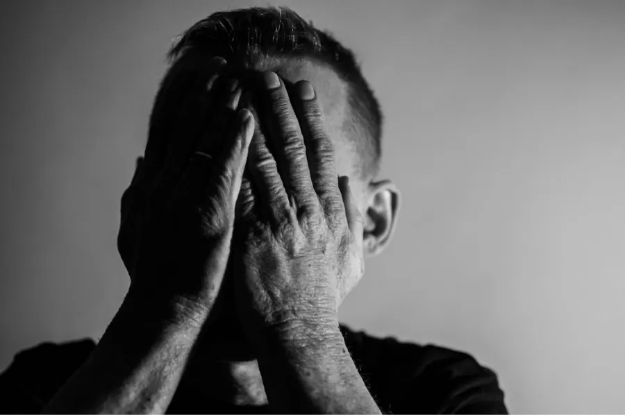 Black and white photo of a short haired person covering their face with their hands on a gray background. Hands are weathered and no portion of the face is visible.