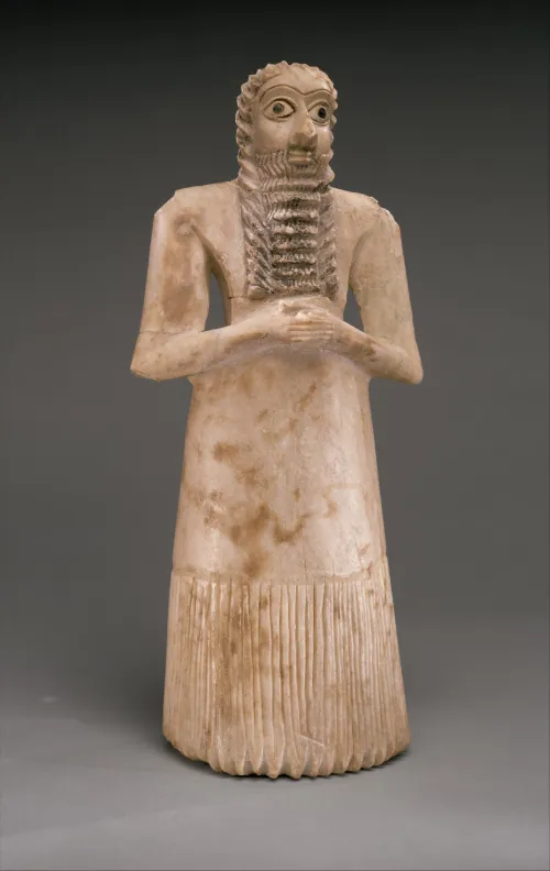 A brown and white statue of a man is shown on a gray background. He wears long robes with pleats along the bottom third and has long wavy hair that runs into his long, rectangular, wavy beard. His eyes are very large and almond-shaped with big black pupils and high eye brows. His nose is large and he is looking forward. His hands are clasped together in front of him.