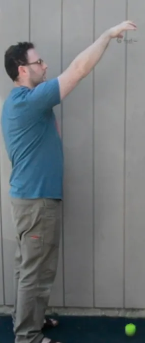 A man in a blue shirt and khaki pants stands sideways  against a gray paneled wall, arm extended upward, and has dropped a yellow tennis ball. The ball is on the ground.