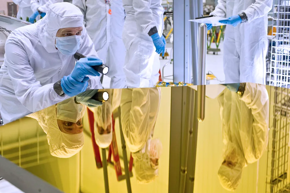 People in white clothing covered from head to toe and wearing blue colored gloves are working in a research laboratory setting, one person holding a flash light and analyzing and another reading a manuscript and so on. Their images are seen on a smooth colored glass top of a work table.