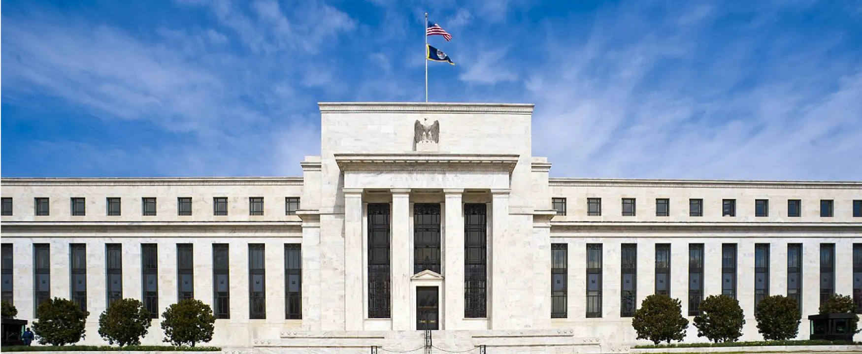 The Marriner S. Eccles Federal Reserve Board Building is shown.  The very large building, located in Washington DC, is constructed of white marble and has a grand entranceway.