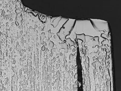 A metallic surface at high magnification appears textured with hundreds of irregularly shaped pits and variations. At one location to the right of the center, a very obvious dark groove appears, rising from the bottom and tapering to a point toward the top.