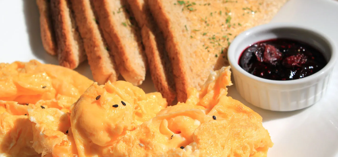 This is a photo of a plate with a large pile of eggs in the foreground and six slices of toast in the background. There is a small dish of red jam sitting near the toast on the plate.