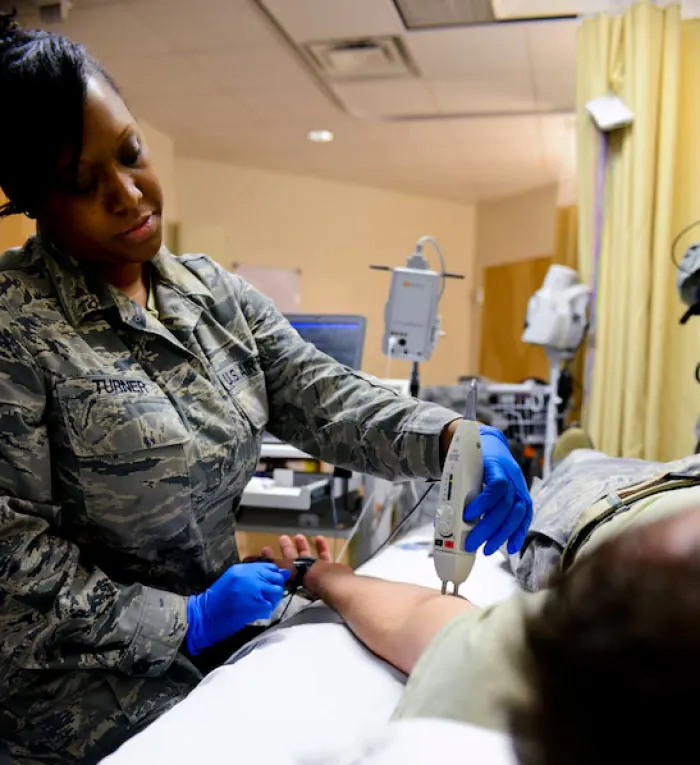 The image shows a medical professional in a military uniform performing a nerve conduction study (NCS) on a patient’s arm. The professional, wearing blue gloves, is holding a handheld device against the patient’s forearm to measure nerve response. The patient is lying on a bed, with their arm extended and connected to electrodes. Medical equipment and monitors are visible in the background, indicating a clinical setting. The focus is on the interaction between the medical professional and the patient, highlighting the procedure being performed to assess nerve function.
