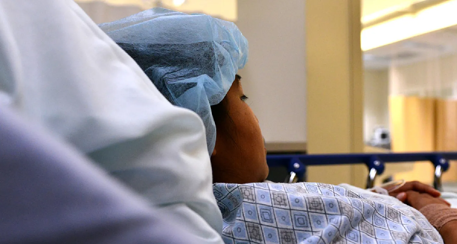 Photo of individual lying in a hospital bed in a gown and hair net.