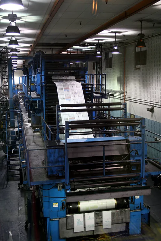 A printing press fills a long room, rolling out large sheets of printed newspaper.