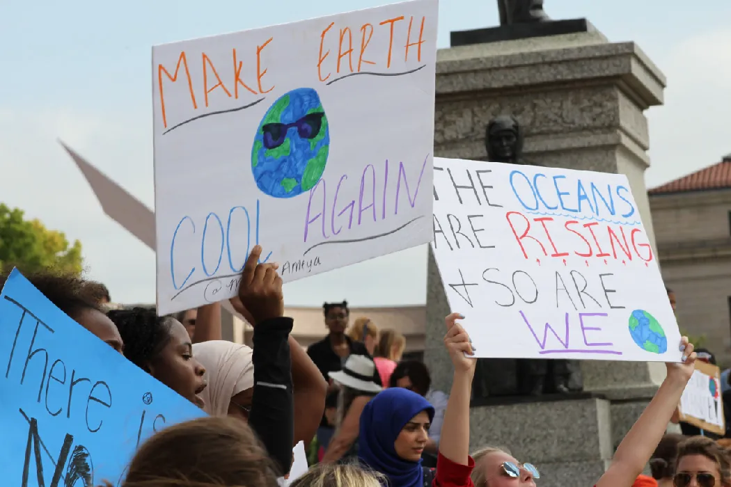 A large group of demonstrators stand outside holding hand-lettered signs that say Make Earth Cool Again and The Oceans Are Rising And So Are We.