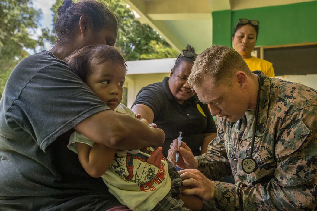 A person in military fatigues injects a needle into the thigh of a small child. The child sits on an adult’s lap while two other adults look on in the background.