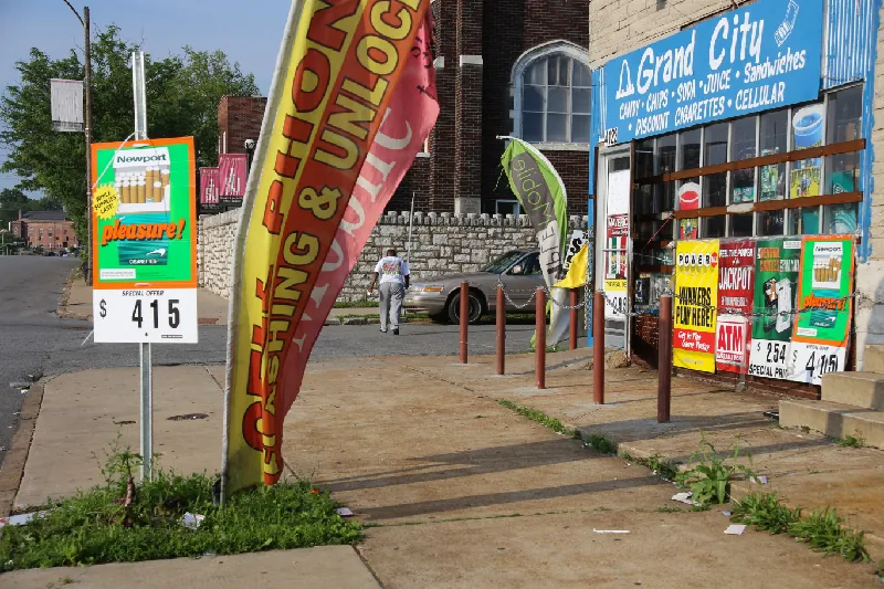 The sign on a store on the corner of a city block reads “Grand City. Candy. Chips. Soda. Sandwiches. Discount Cigarettes. Cellular.” Advertisements for cigarettes and lottery games are on the window and in front of the store.