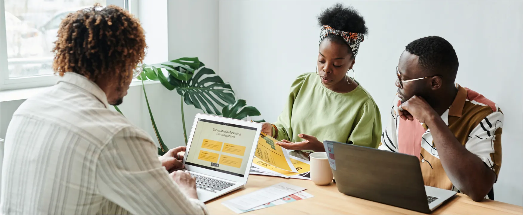 A woman and two men sit at a table looking at laptops and paperwork talking to each other.
