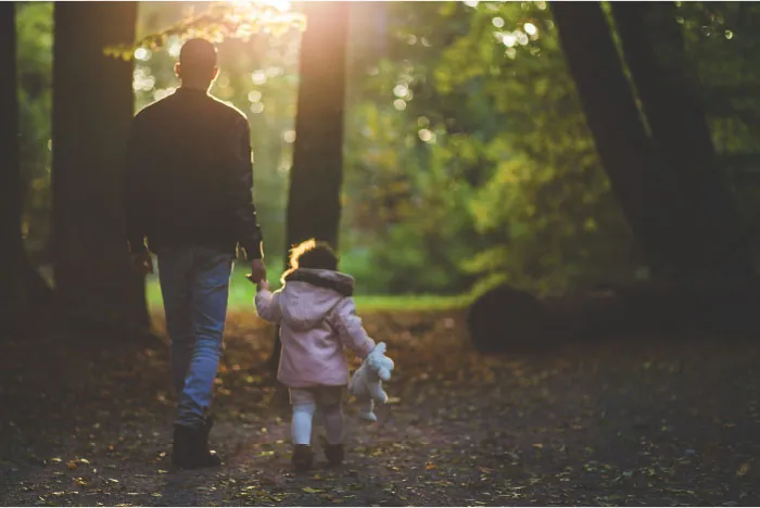 Adult and child with stuffed animal walk through forest holding hands.