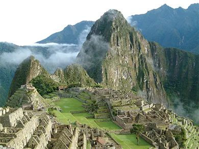 A photograph of Machu Picchu shows the ruins of a complex of buildings with stone walls, stepped terraces green with grass, and a pyramid, with high mountains in the background.
