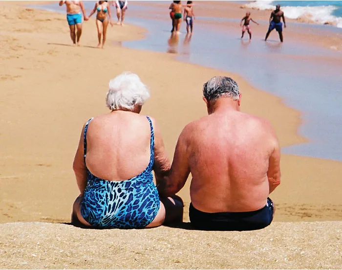Two older adults are shown sitting on a beach in bathing suits, exposing their skin to the sun.