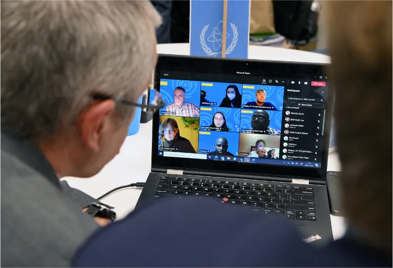 Two people are looking at a laptop screen that displays eight people’s faces on the screen. At the right is a list of participants who have dialed in to the meeting.