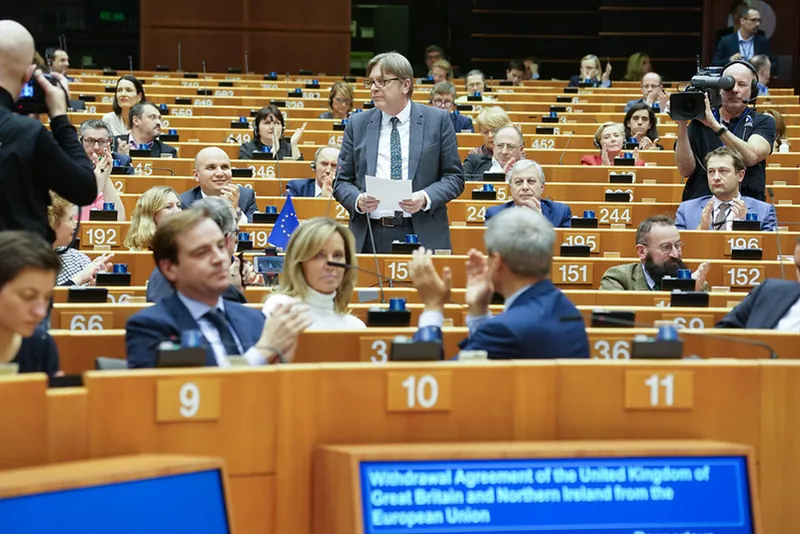 A legislator, surrounded by a photographer, a cameraperson, and many seated colleagues--some of whom applaud, stands holding a piece of paper at one of many connected, numbered wooden desks arranged in tiered rows.