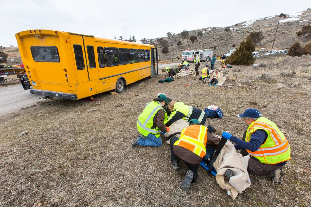 EMS workers wearing reflective vests gather outside in small groups near a yellow school bus. In the foreground, several kneel in the grass next to a dummy on a backboard. Other emergency vehicles are visible in the background.