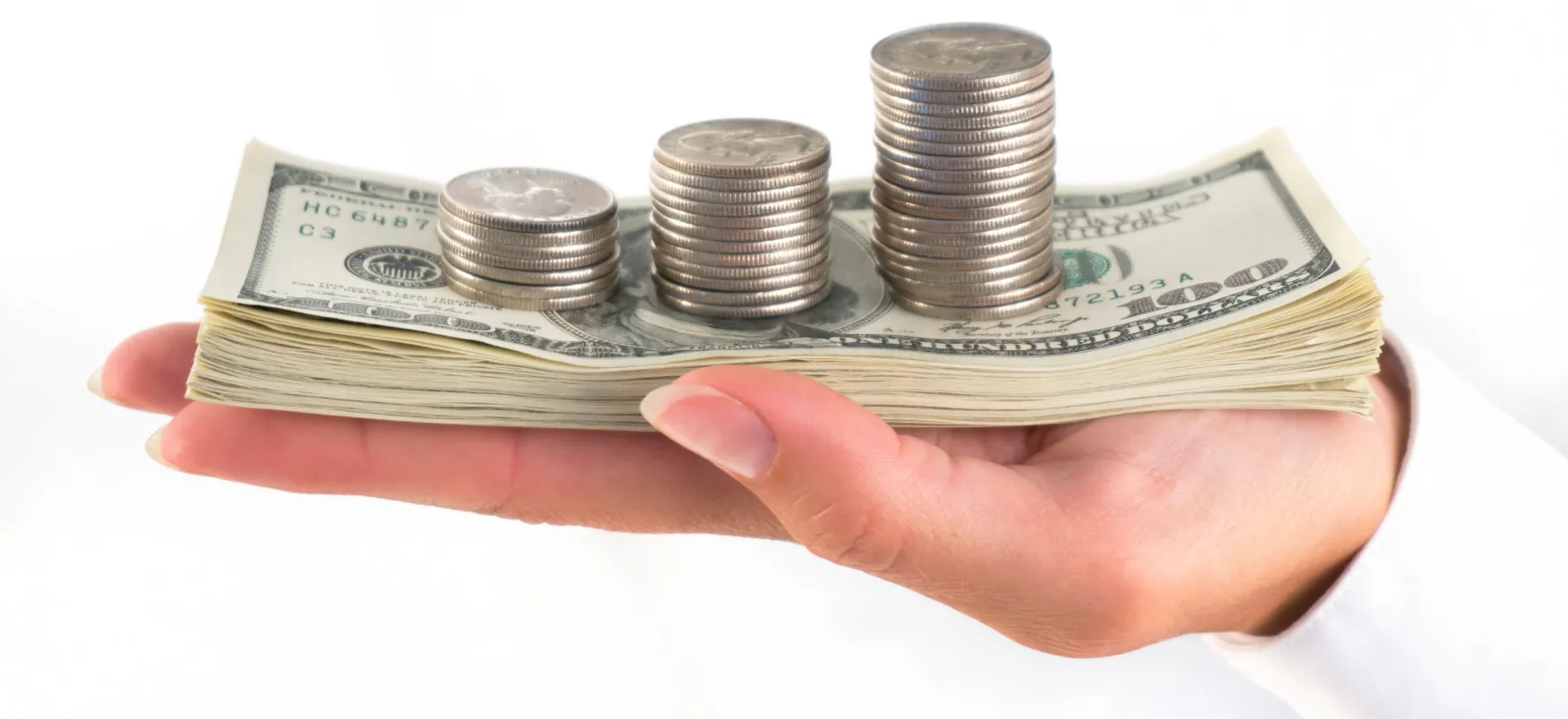 A hand holds a stack of $100 bills with three columns of coins stacked on top of the bills.