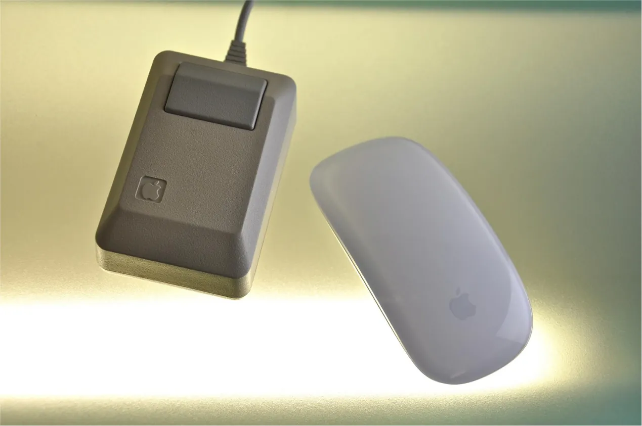 Two computer mouse devices from Apple are pictured side by side. The older one is a rectangle with a button and a wire.  The newer one is oval and much more smooth, with no buttons and no wires.