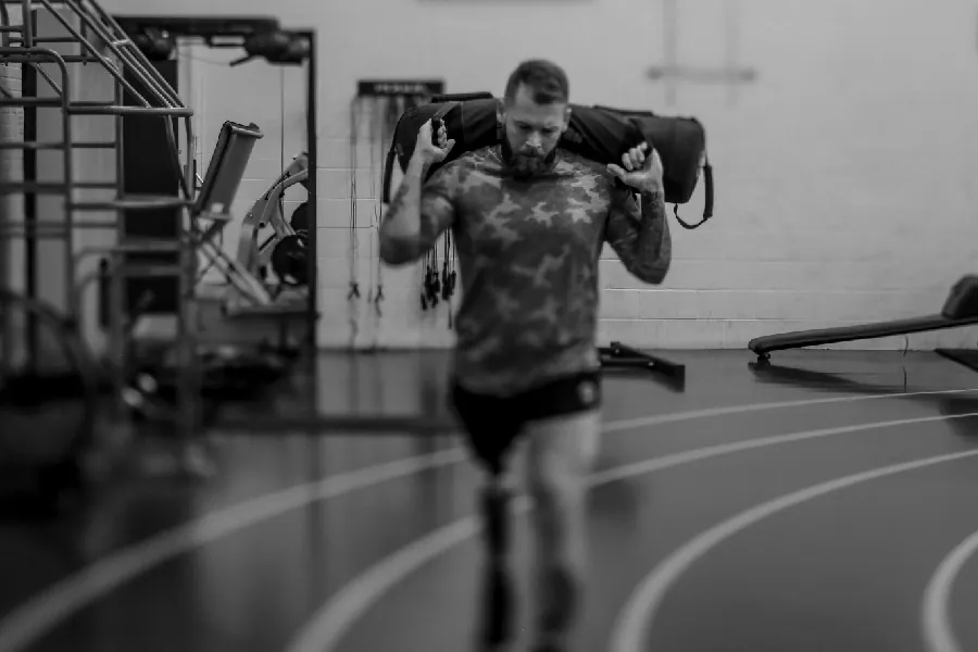 A person with a prosthetic leg walks around an indoor track, carrying a long duffle bag on their shoulders.