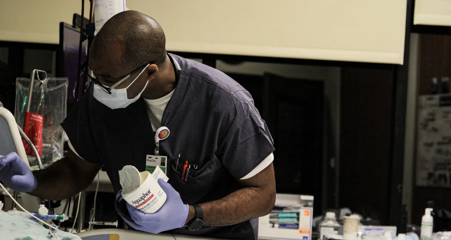 A color photograph of a nurse standing at the bedside putting ointment to a patient.