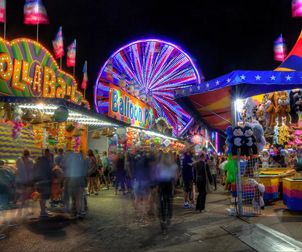 A vibrant nighttime carnival scene with colorful lights, a glowing Ferris wheel, game booths like Balloon Pop, hanging prizes, and a crowd of people enjoying the festive atmosphere.