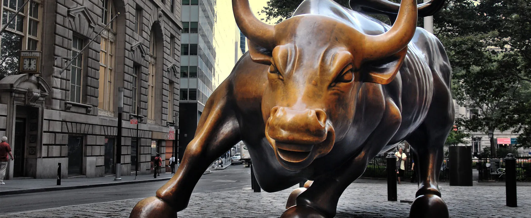 A picture of the bronze statue Charging Bull by Arturo di Modica. The statue, located on a cobblestone sidewalk in New York City’s Wall Street area, is approximately eleven feet tall and sixteen feet long, and depicts an a bull in an aggressive stance, as if it is in the process of running forward.
