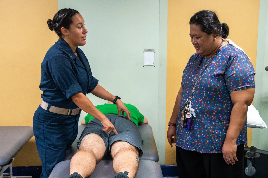 A client lies on their stomach on a therapy table. Two therapists stand on opposite sides of the table, discussing the client’s care.