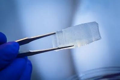 A person uses forceps to hold a semi-transparent three dimensional rectangle, which is about 4 centimeters long and about a centimeter wide. The surface of the object is grooved and textured.