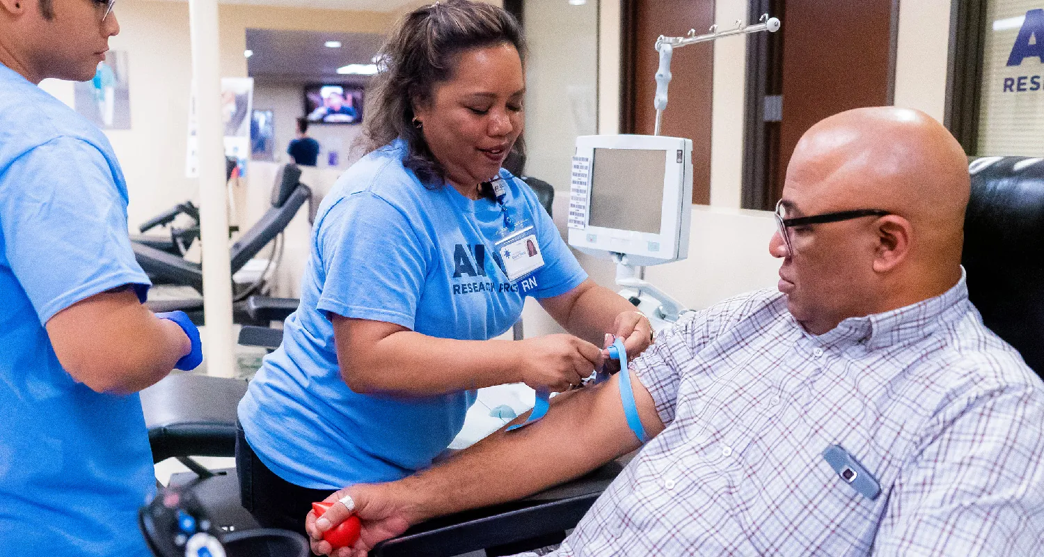 A client sits in a chair with their right arm extended on an armrest. A nurse ties a tourniquet onto their arm above the elbow.