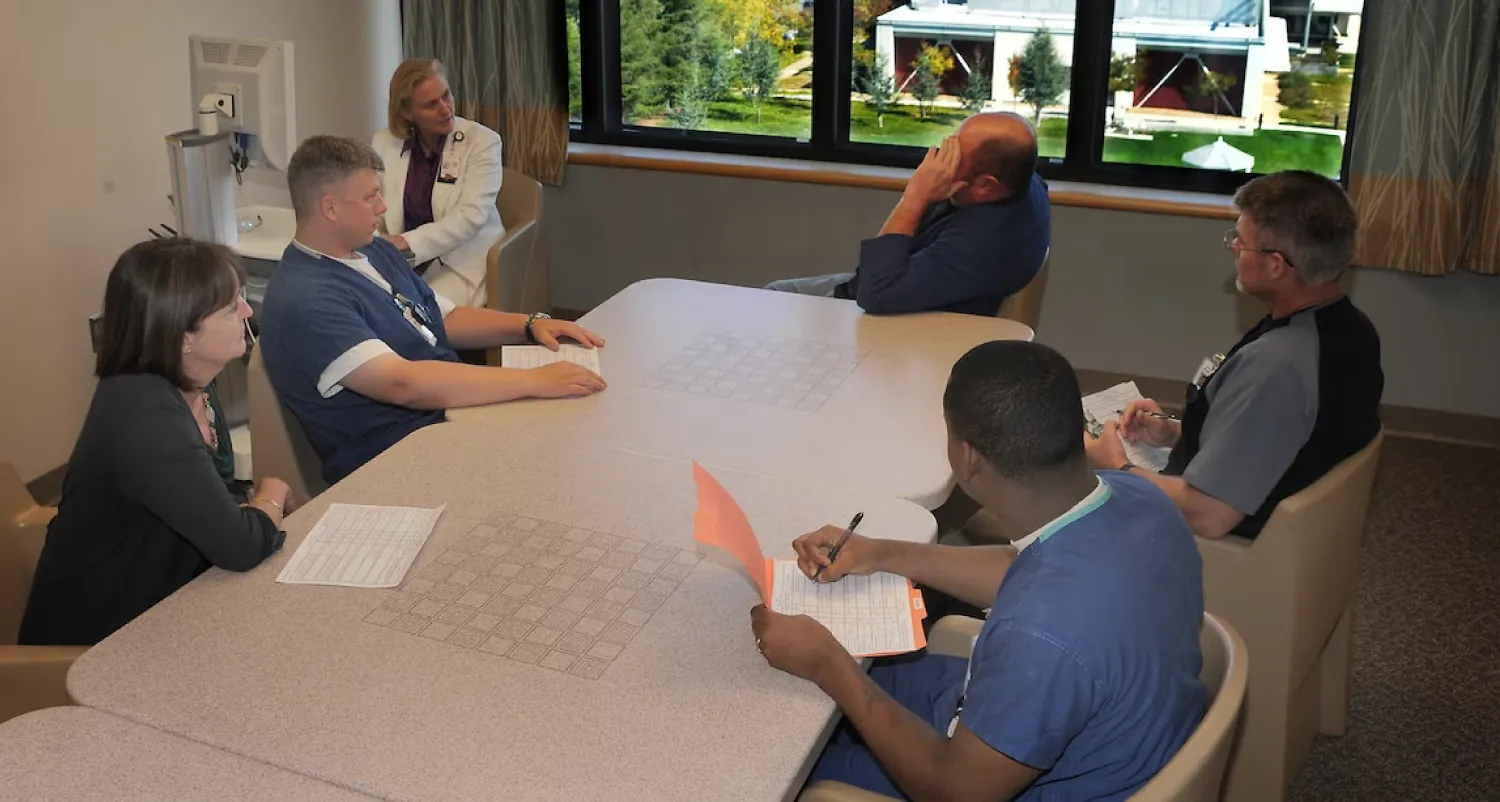 A color photograph of a group of health-care providers sitting around a table looking at a client holding his head.