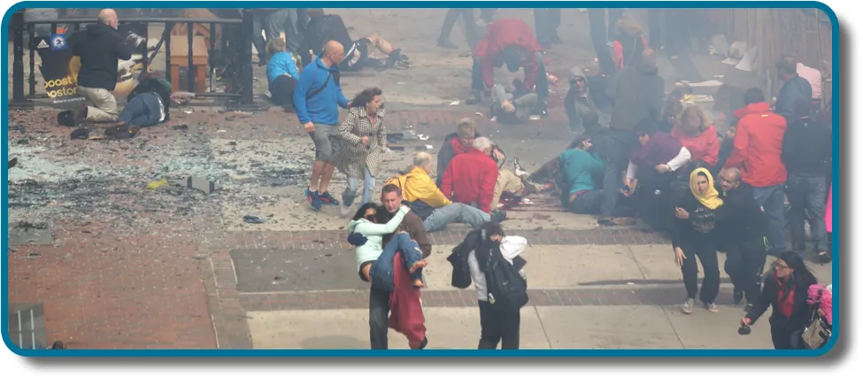 A photograph shows a crowd  at the site of the Boston Marathon bombing immediately after it occurred. Debris is scattered on the ground, several people appear to be injured, and several people are helping others.