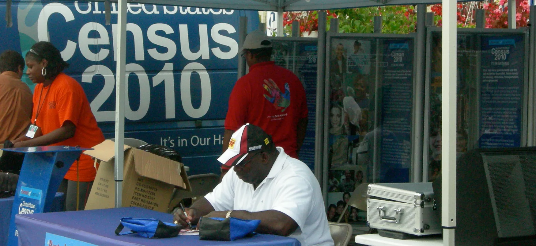 A person fills out paperwork at a table under a canopy with “Census 2010” signs, while a diverse team assists.