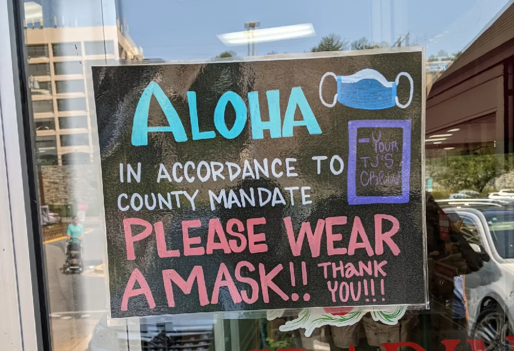 A sign in a window states: Aloha. In accordance to community mandate please wear a mask!! Thank you!!!  A drawing of a paper surgical mask appears in the corner of the sign.