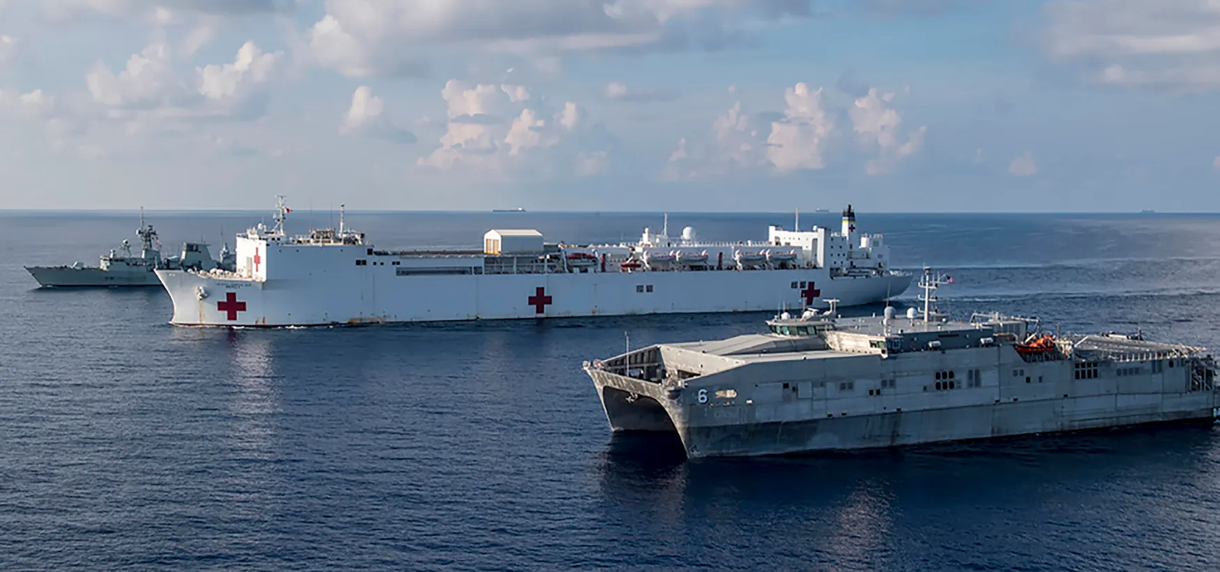 Three navy ships sail in formation. In the foreground is a transport and equipment vessel with a large opening underneath for loading and unloading. Next to it is a United States hospital ship, clearly marked with red crosses on a white hull. Next to it is another military vessel with clearly visible antenna and armorments.