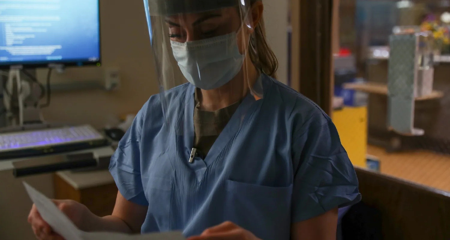 A nurse wearing a mask and a face shield reviews a patient’s file.