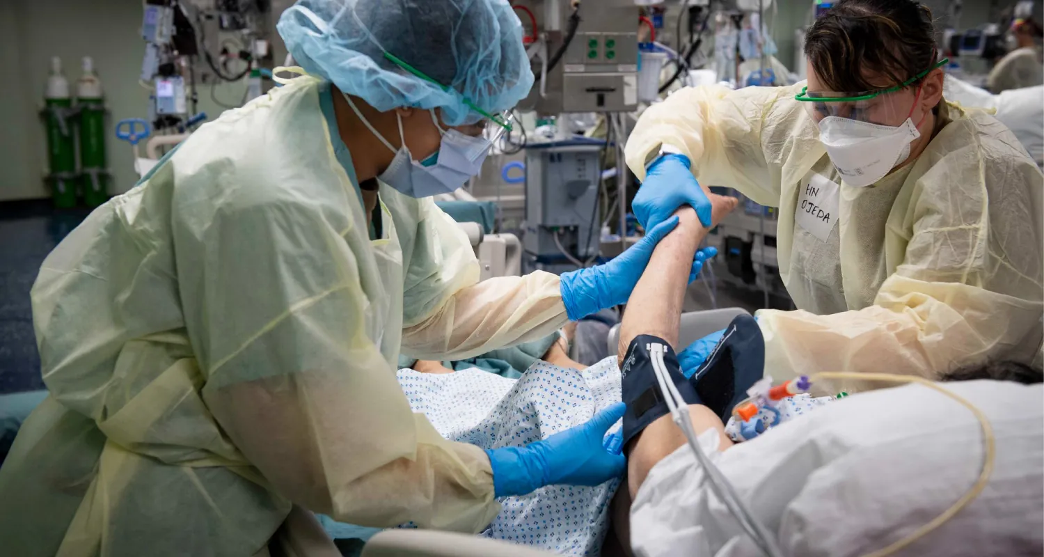 The image shows healthcare workers in full protective gear treating a patient in a hospital bed. One worker holds the patient’s arm, while another adjusts equipment. The patient is connected to tubes and monitors in a critical care setting, with medical equipment visible in the background.