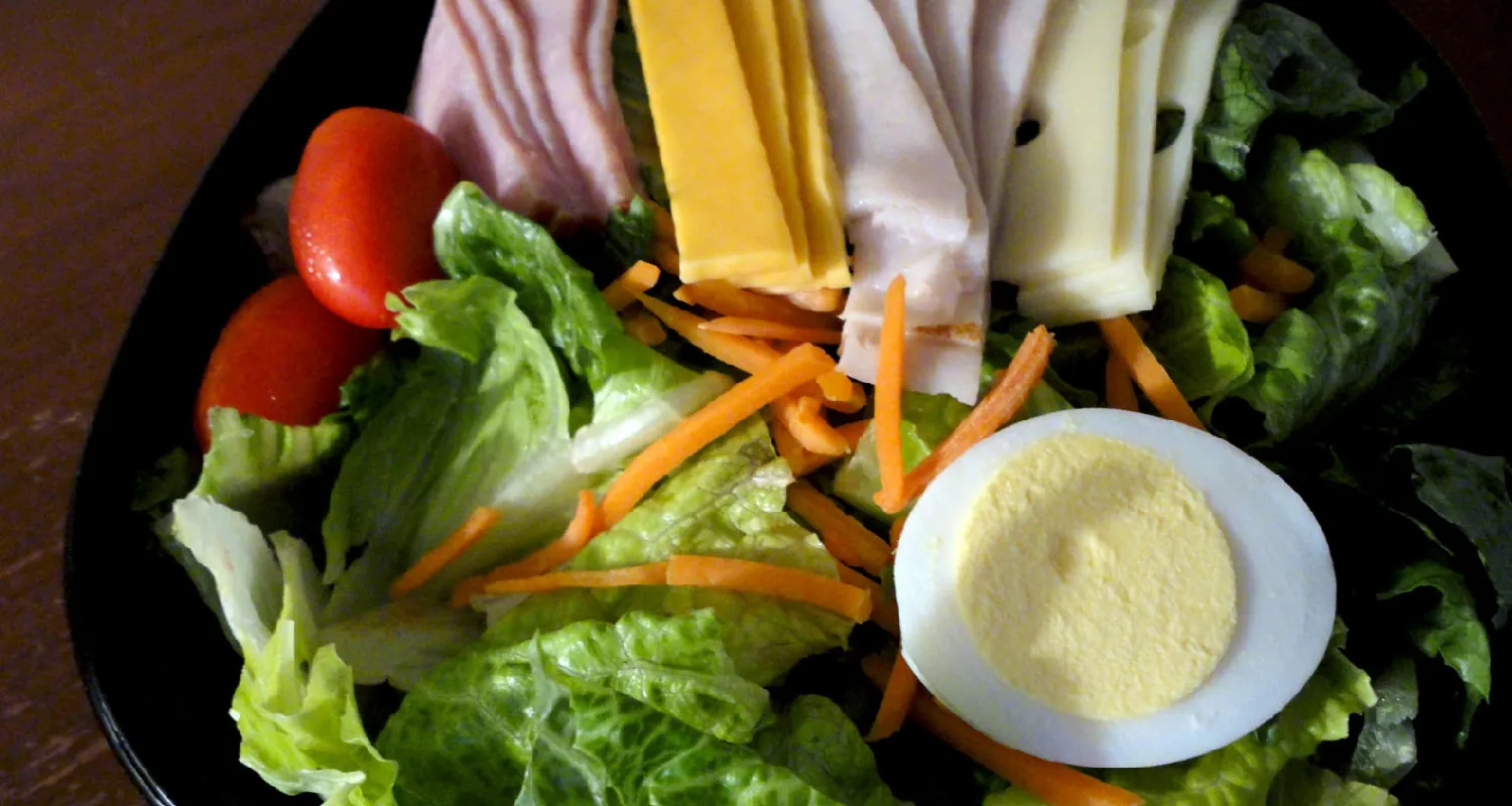A chef salad consisting of lettuce, cherry tomatoes, carrots, cheese, deli meats, and a sliced hard-boiled egg is arranged on a plate.