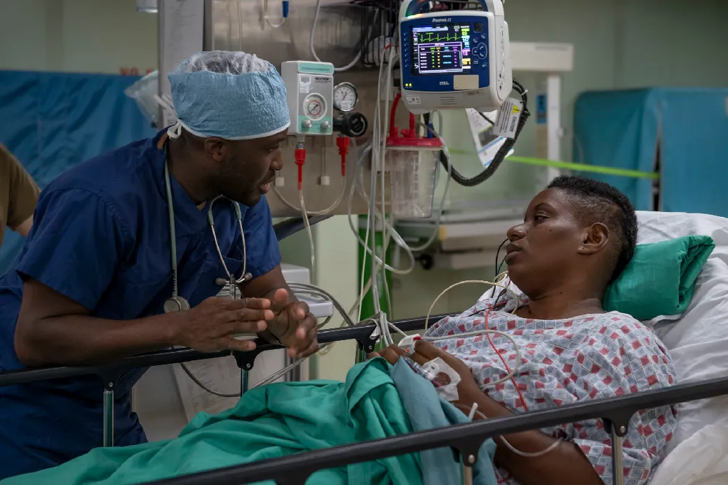 A nurse dressed in scrubs speaks with a client who lays listening in a hospital bed.
