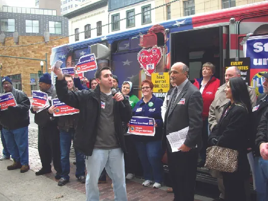 An image of a group of people standing in front of a bus. Some of the people hold signs.
