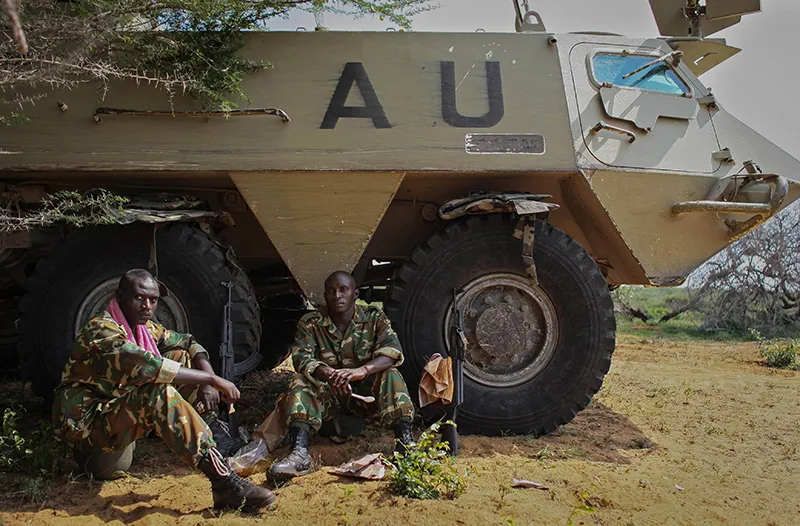 Two soldiers dressed in camouflage sit on the ground beside a tank with the letters A U painted on the side.