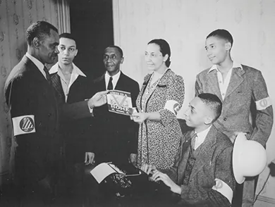 A photograph shows five Black men and a Black woman participating in the Double V campaign. A young man sits at a typewriter, and the woman hands a man a pamphlet, the cover of which reads “This is a [Double V insignia] Home.” All wear armbands.