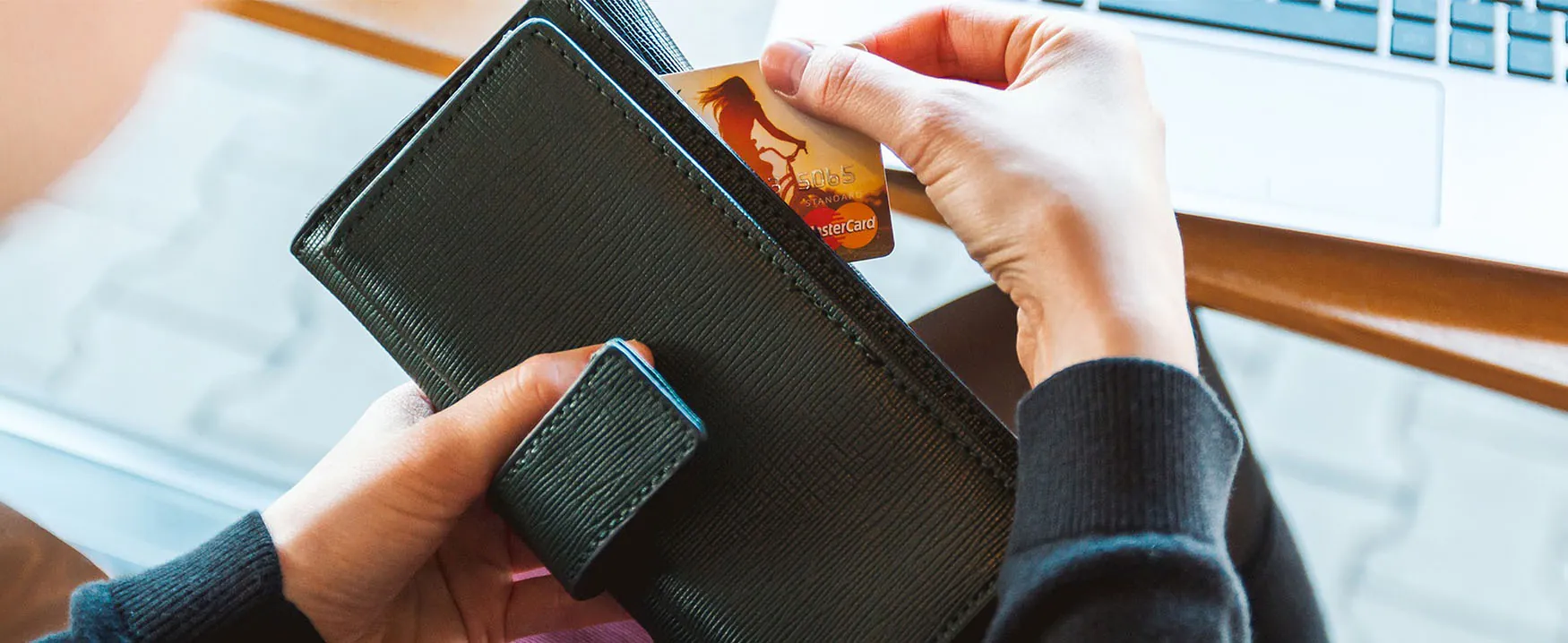 A picture of a person removing a credit card from their wallet. A laptop computer is in the background.