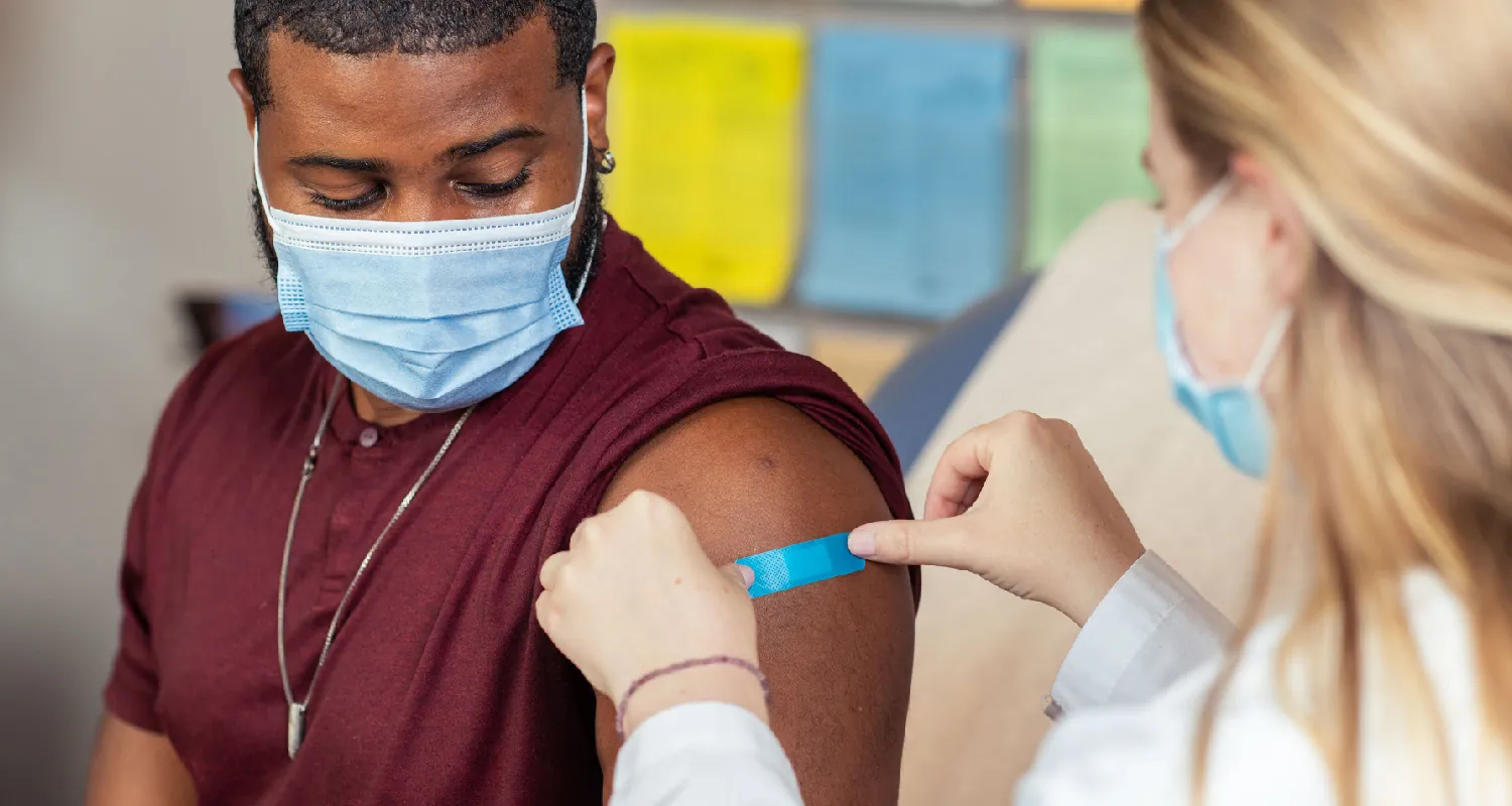 Two seated people wear surgical masks. The person on the left wears a t-shirt with the sleeve rolled up, and the person on the right places a small bandage on their upper arm.