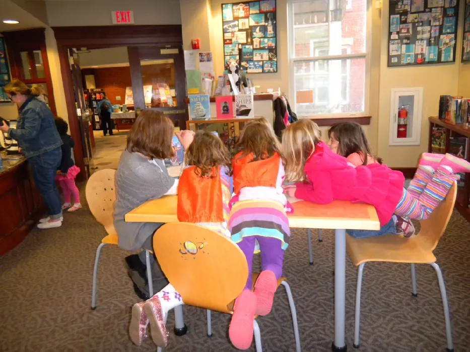 A group of children sit around a table at a library, leaning forward and listening to an adult reading a book.