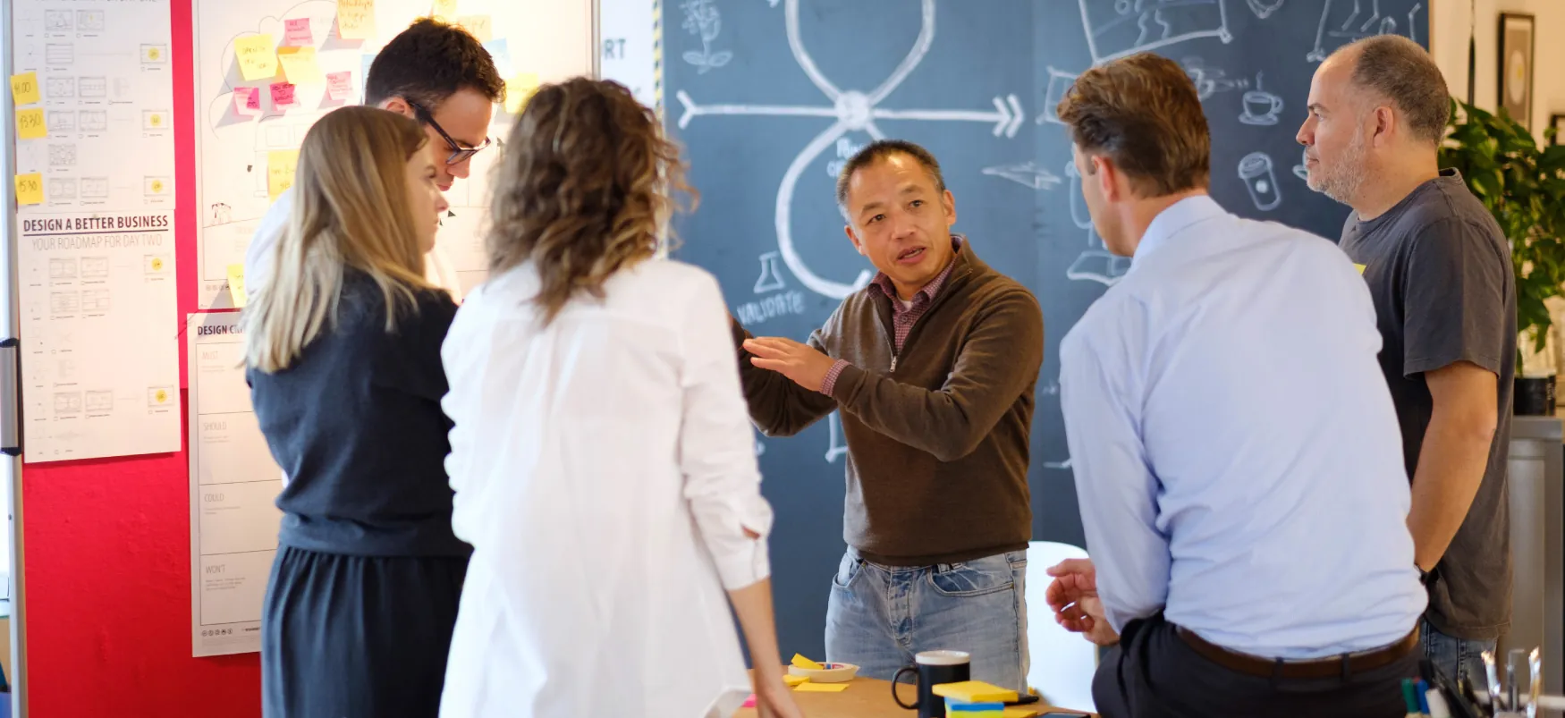 Grupa osób stoi w kręgu i rozmawia przed tablicą z narysowanymi na niej diagramami.